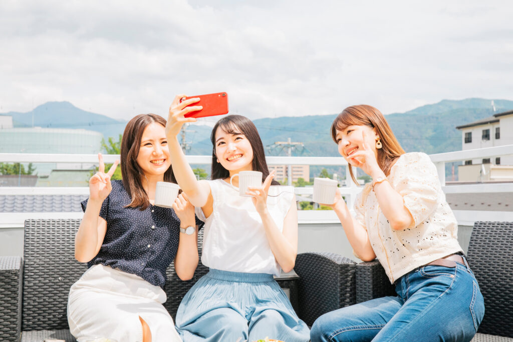 屋上でコーヒーカップ片手にピースサインをして自撮りをしている女性3人